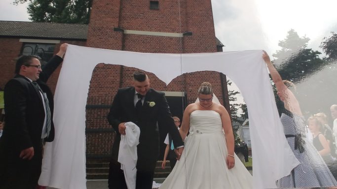 Brautpaar und Gäste halten einen weißen Stoffbogen vor einer Kirche.
